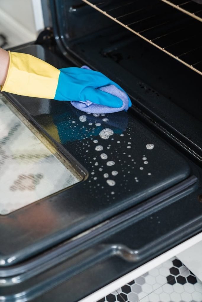Close-up of cleaning an oven door with gloves and detergent in a kitchen.