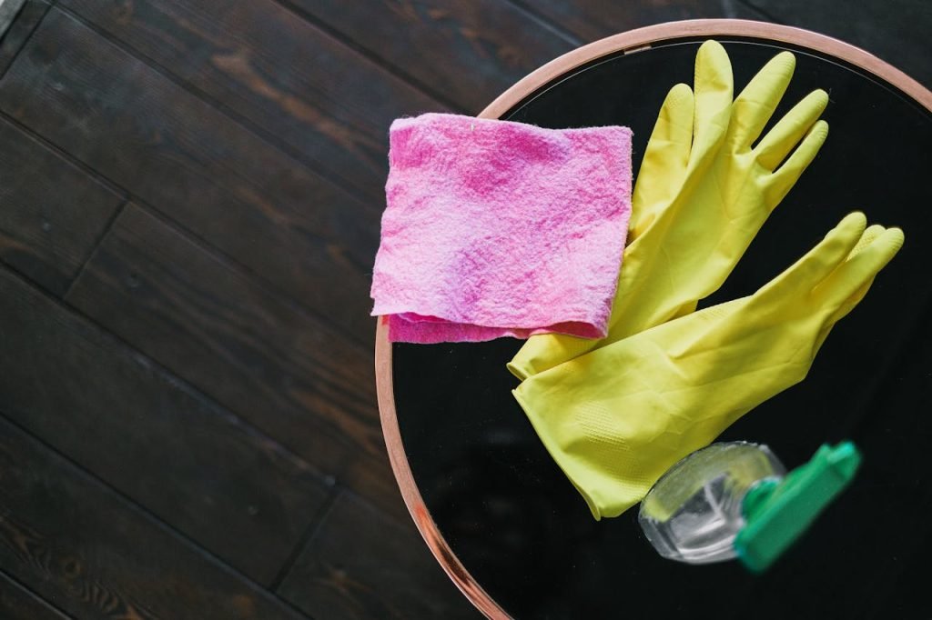 Yellow gloves and pink cloth on a table with cleaner bottle, viewed from above.